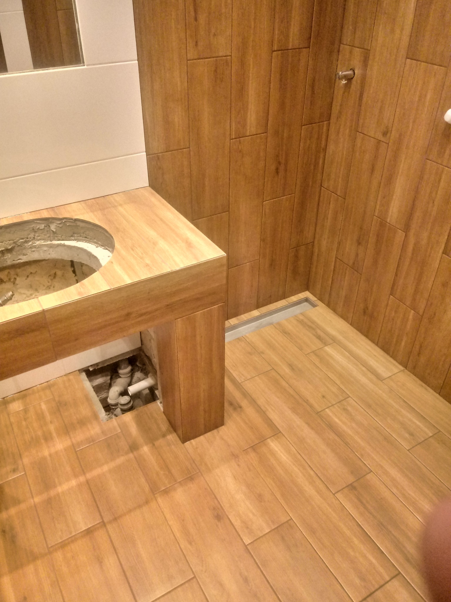 Łazienka w trakcie remontu, widoczne drewnopodobne płytki na ścianach i podłodze, niedokończona zabudowa umywalki z widocznymi rurami i otworem na zlew.