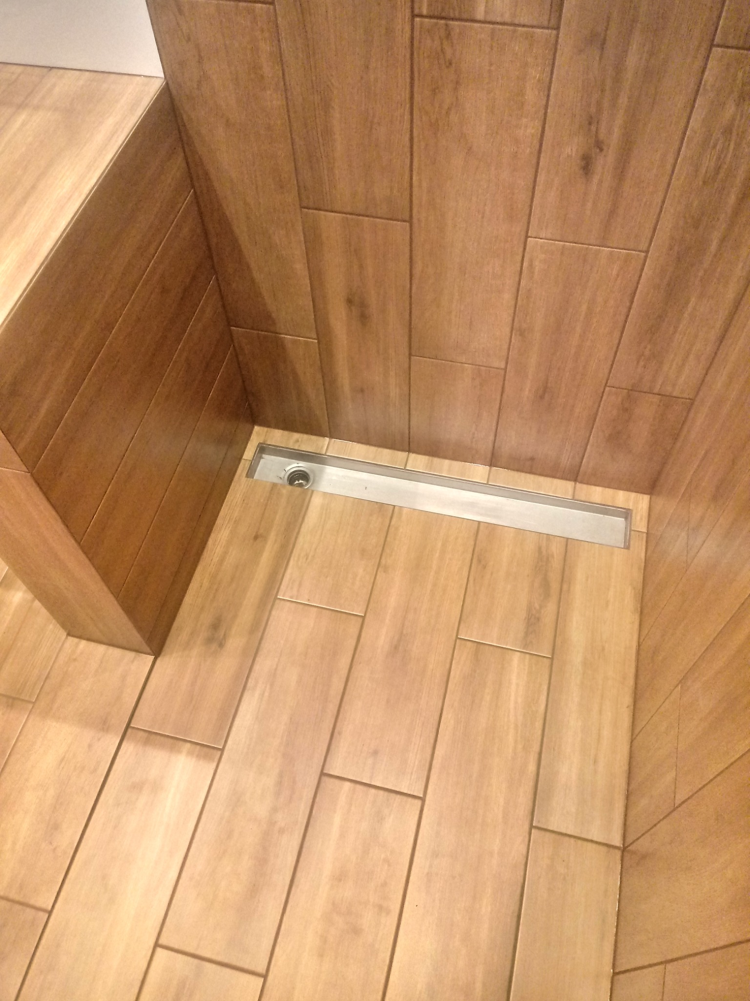 Wykończona kabina prysznicowa z odpływem liniowym i okładziną z płytek imitujących drewno, ułożonych pionowo.