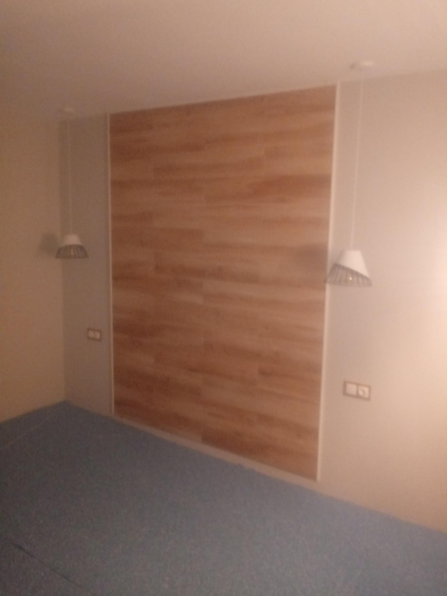 Ściana wyłożona panelami imitującymi drewno, z wiszącymi lampami po bokach i szarą podłogą.