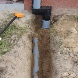 Wykop z ułożonym szarym rurociągiem kanalizacyjnym w ziemi, połączonym z pionową rurą spustową z dachu budynku, widoczny czarny osadnik z kratką, obok leży pomarańczowa łopata.