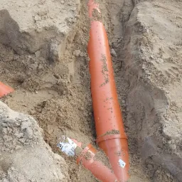 Pomarańczowe rury kanalizacyjne zakopane w piasku, widoczne złącza i fragment etykiety na rurze. W tle widoczne narzędzia pracy.