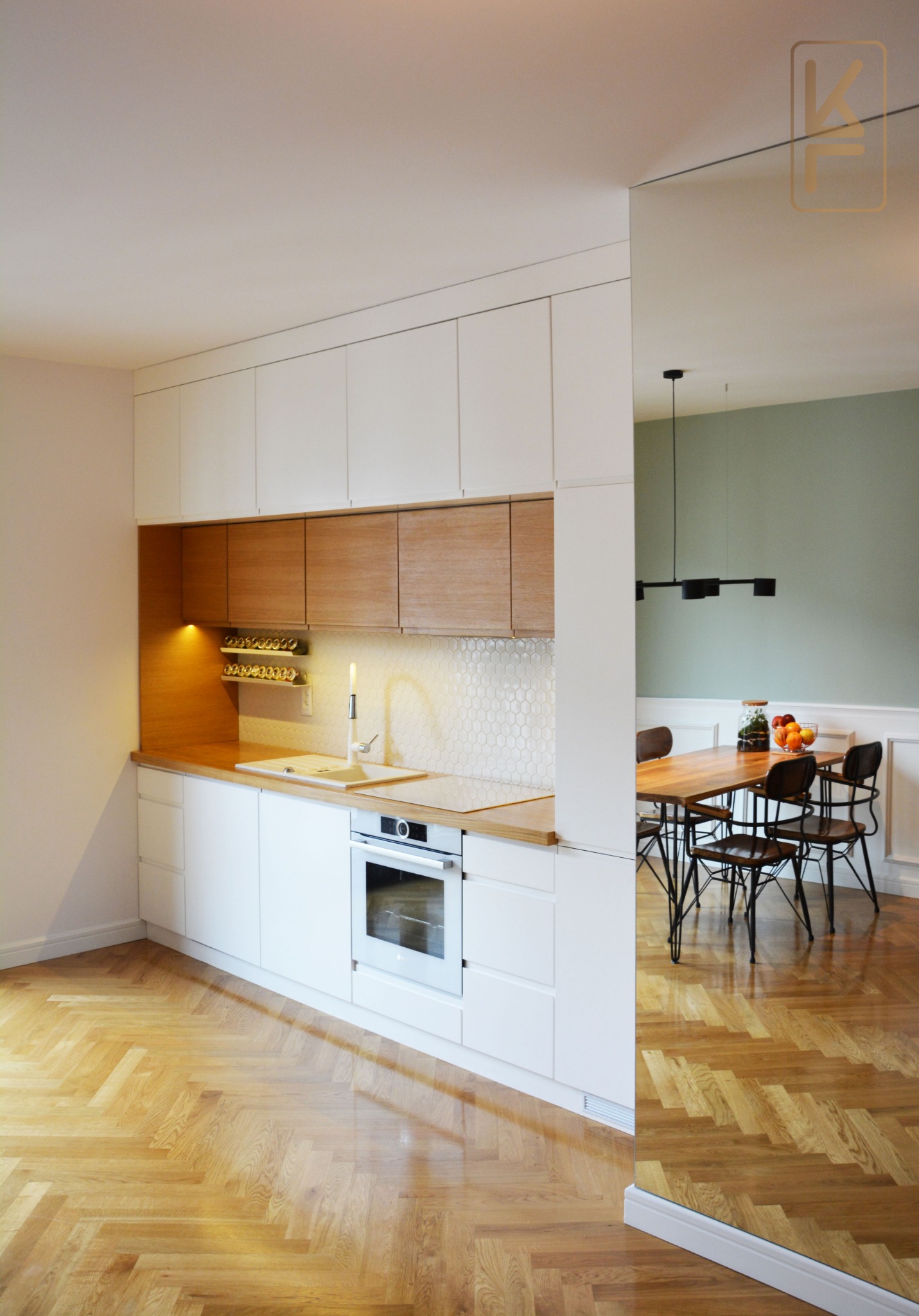 Nowoczesna kuchnia z białymi szafkami i drewnianymi akcentami, widoczny stół z krzesłami w tle, parkiet ułożony w jodełkę.