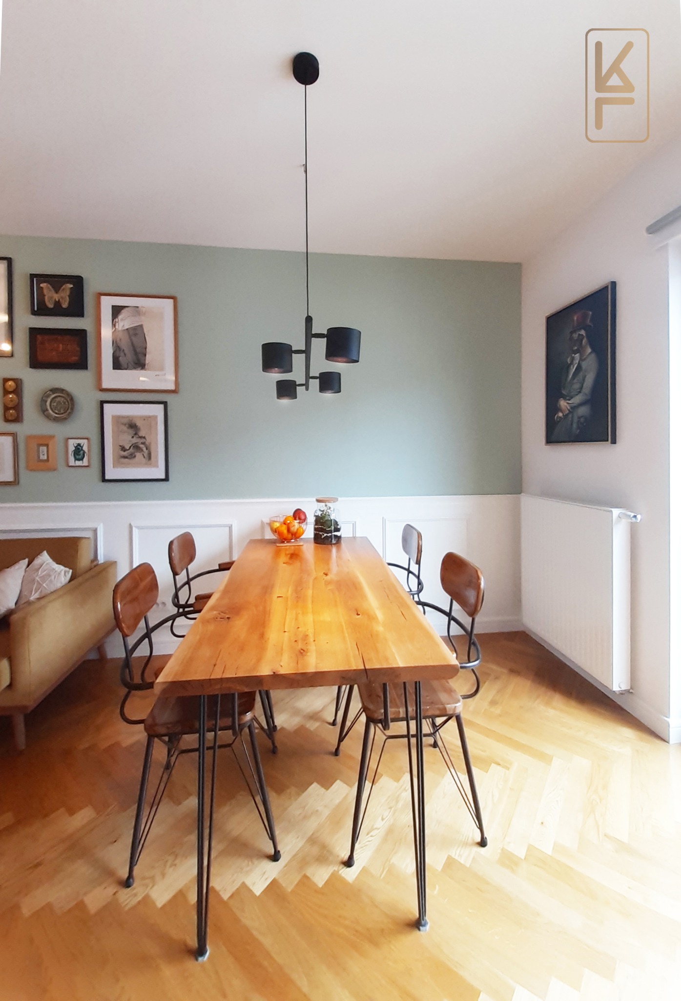 Jasne wnętrze jadalni z drewnianym stołem, krzesłami na metalowych nogach, parkietem w jodełkę i ścianą w kolorze pistacjowym z galerią obrazków.