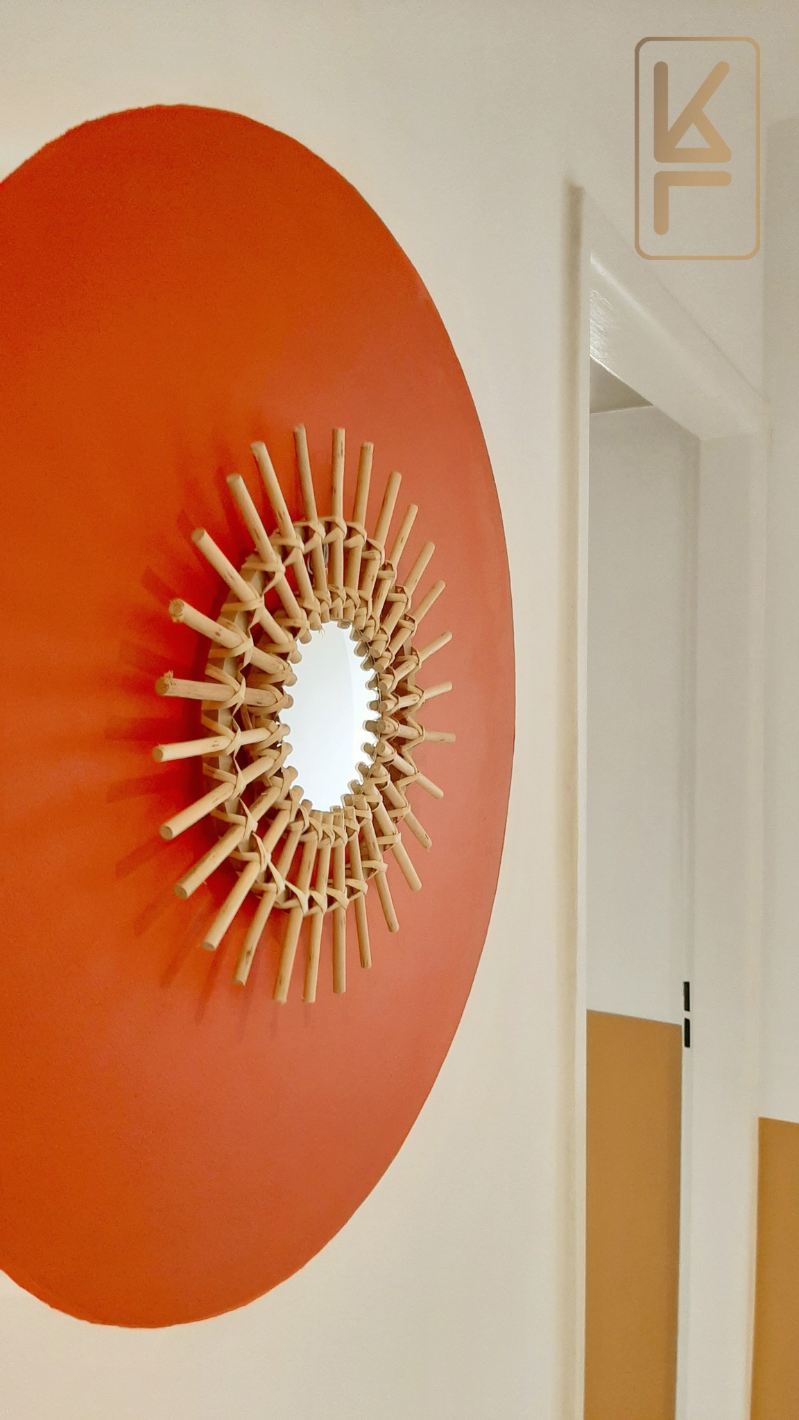 Okrągłe lustro w ramie z naturalnego rattanu, umieszczone na pomarańczowym okręgu na kremowej ścianie, widoczne z boku, z fragmentem białej futryny drzwi w tle.