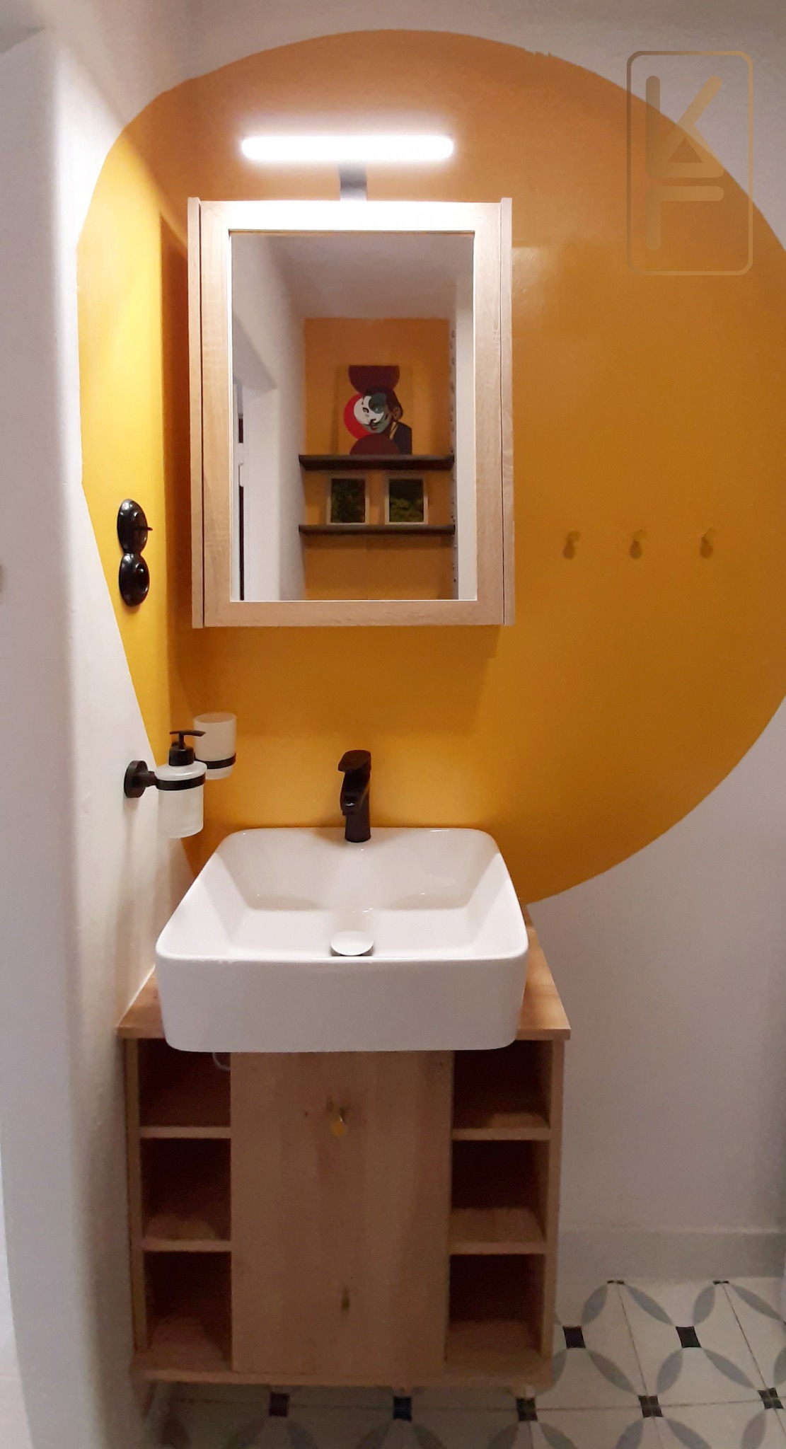 Nowoczesna łazienka z umywalką nablatową, szafką z otwartymi półkami, lustrem z oświetleniem i żółtą ścianą z abstrakcyjnym motywem koła oraz czarnymi akcentami w postaci baterii i akcesoriów.