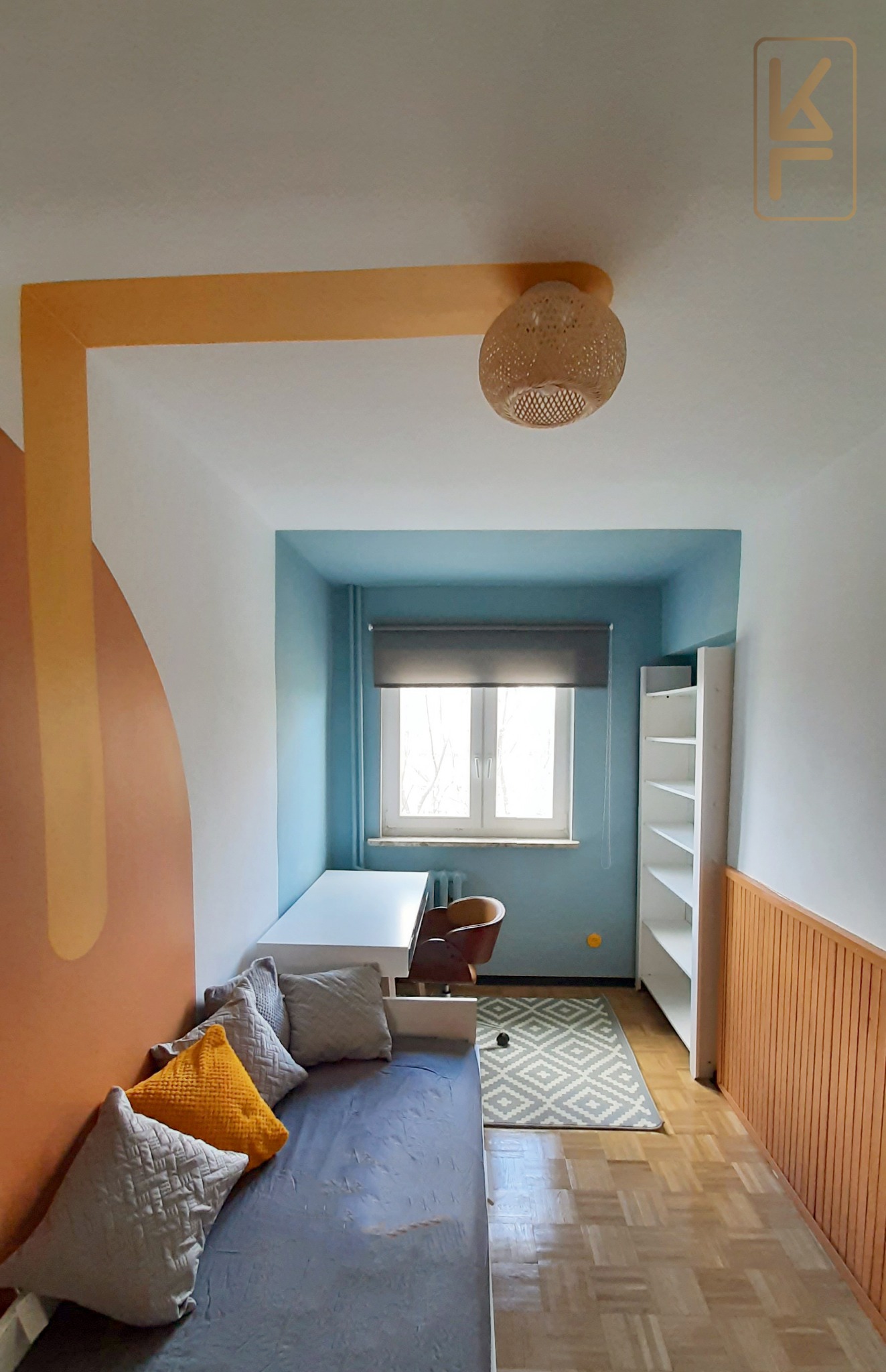 Pokój młodzieżowy z pomalowaną ścianą w odcieniach brązu i żółci, biurkiem pod oknem, łóżkiem z szarymi i żółtą poduszką oraz plecioną lampą wiszącą.