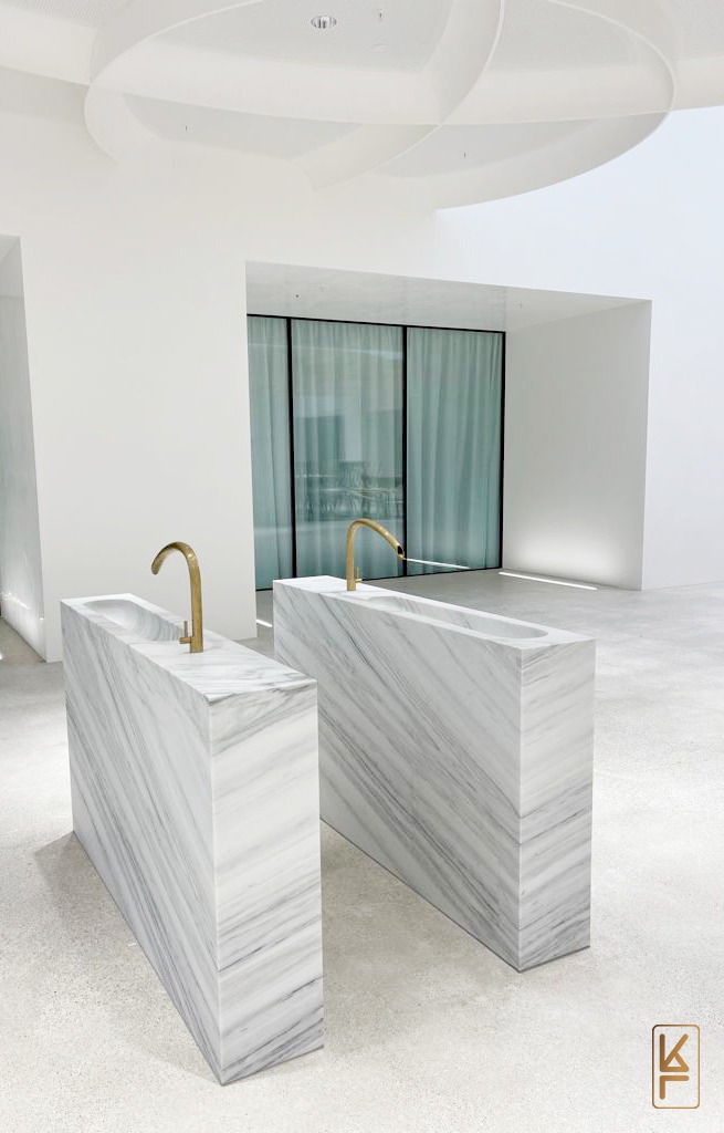 Dwie marmurowe umywalki w minimalistycznym wnętrzu z białymi ścianami i sufitem, złote krany o zakrzywionym kształcie, duże okno z zasłonami w tle.