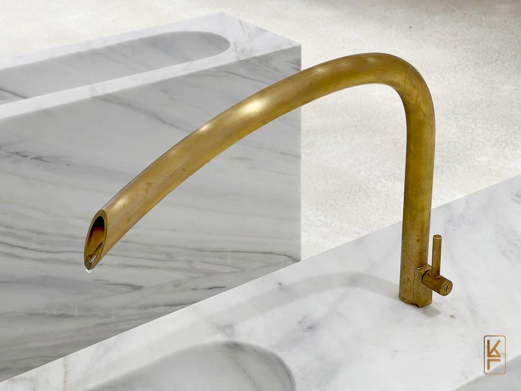 Mosiężna bateria umywalkowa o nietypowym kształcie, z kroplą wody na końcu, zamontowana na blacie z białego marmuru z szarymi żyłami, w tle widoczny fragment umywalki z tego samego marmuru.