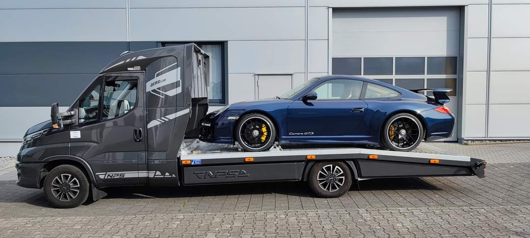 Niebieskie Porsche Carrera GTS na szarej lawecie transportowej, z żółtymi zaciskami hamulcowymi, zaparkowane na kostce brukowej przed szarym budynkiem.