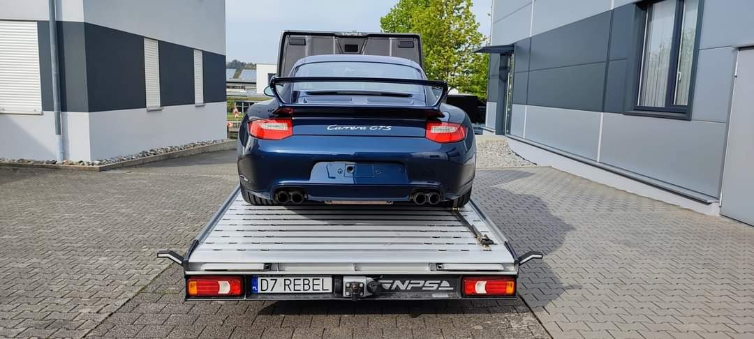 Niebieski Porsche Carrera GTS z otwartą klapą bagażnika, umieszczony na aluminiowej platformie lawety z widoczną tablicą rejestracyjną 'D7 REBEL', na tle nowoczesnego budynku z szaro-białą elewacją.