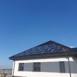 Panele fotowoltaiczne na dachu domu jednorodzinnego z ciemną dachówką, widok z dołu, błękitne niebo w tle.