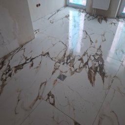 Dream Hause - Położona, błyszcząca, biała podłoga z marmurowym wzorem w łazience. Widoczny odpływ liniowy, grzejnik i okno z niebieską folią ochronną.