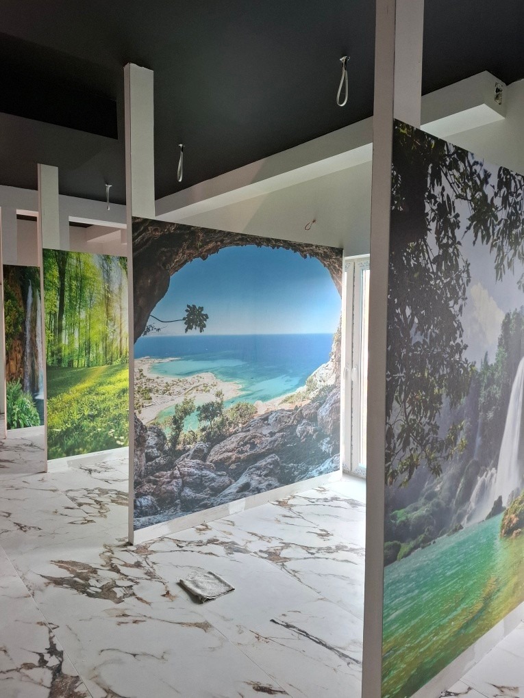 Wnętrze z fototapetami przedstawiającymi krajobrazy: las z wodospadem i morski widok z palmą. Marmurowa podłoga i czarny sufit. Widoczne haki sufitowe.