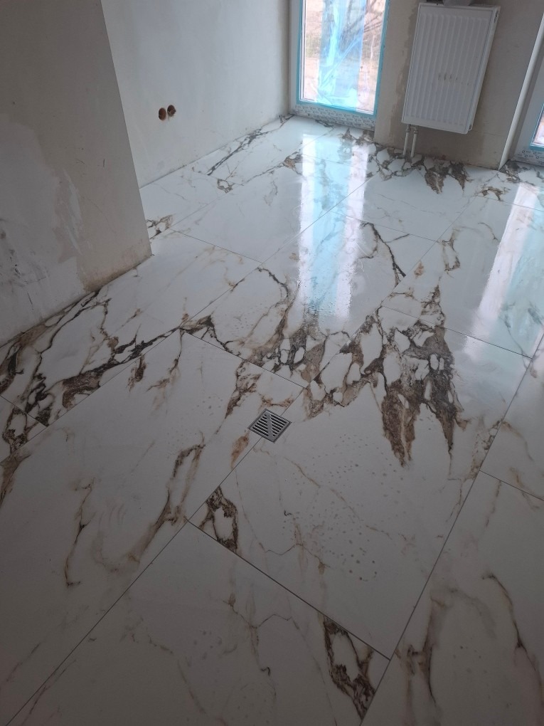 Położona, błyszcząca, biała podłoga z marmurowym wzorem w łazience. Widoczny odpływ liniowy, grzejnik i okno z niebieską folią ochronną.