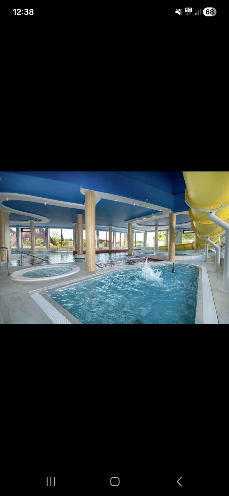Nowoczesny basen kryty z niebieskim sufitem, mozaikowymi kolumnami i żółtą zjeżdżalnią. Woda w basenie lekko wzburzona, widoczne jacuzzi i duże okna.