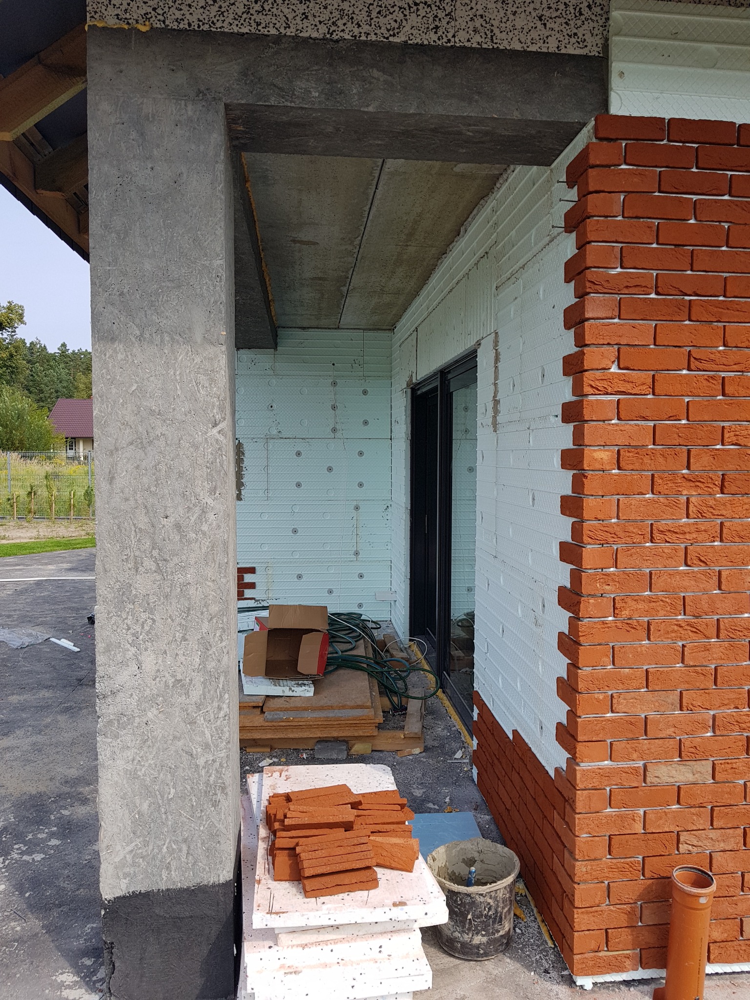 Budynek w trakcie ocieplania i wykańczania elewacji z cegły klinkierowej, widoczna warstwa izolacji ze styropianu, przygotowane cegły, wiadro z zaprawą, betonowy filar zabezpieczony hydroizolacją.