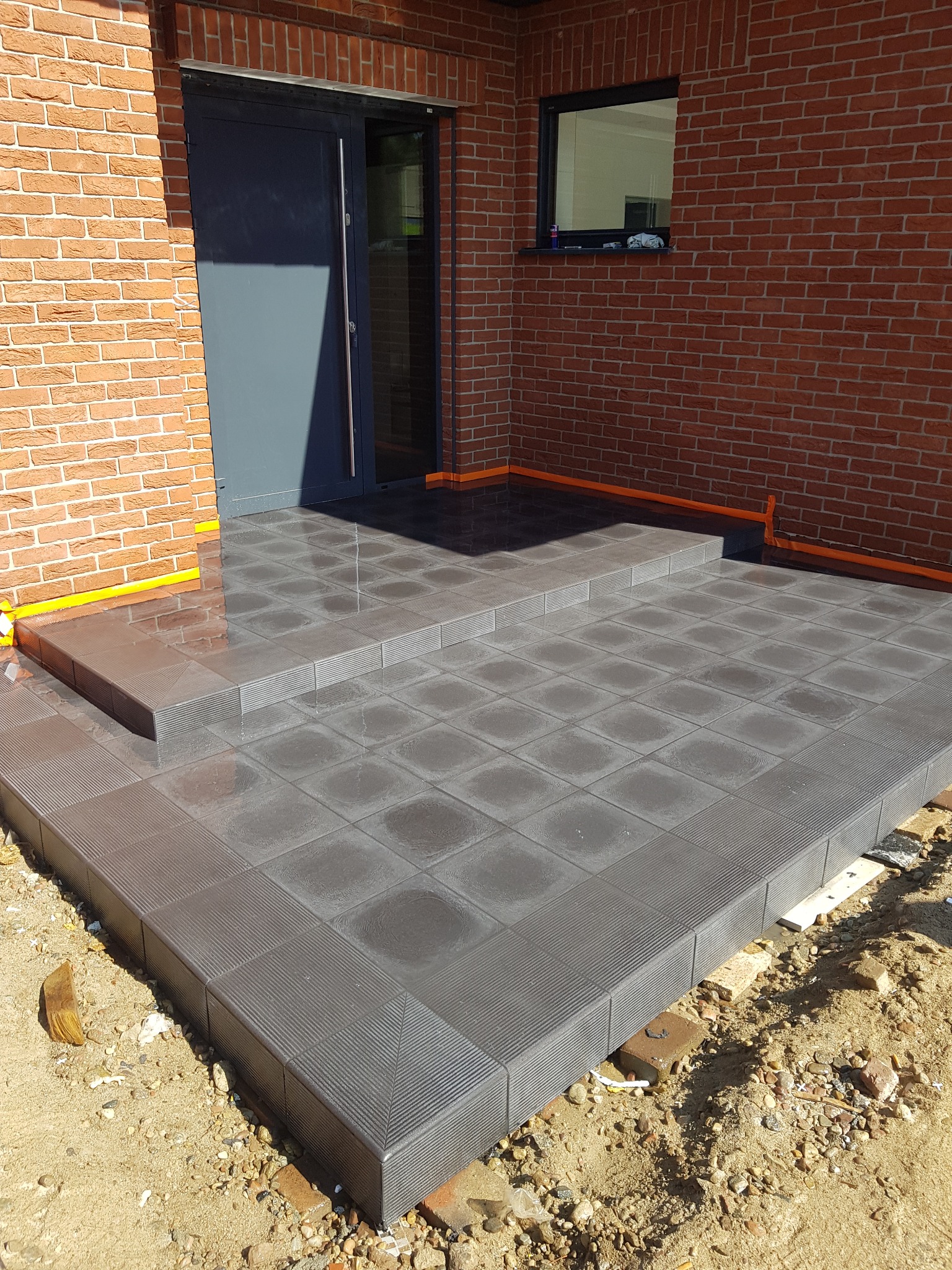 Nowo wyłożony, grafitowy taras z płytek kwadratowych przed wejściem do domu z cegły, zabezpieczony taśmą malarską wzdłuż krawędzi.