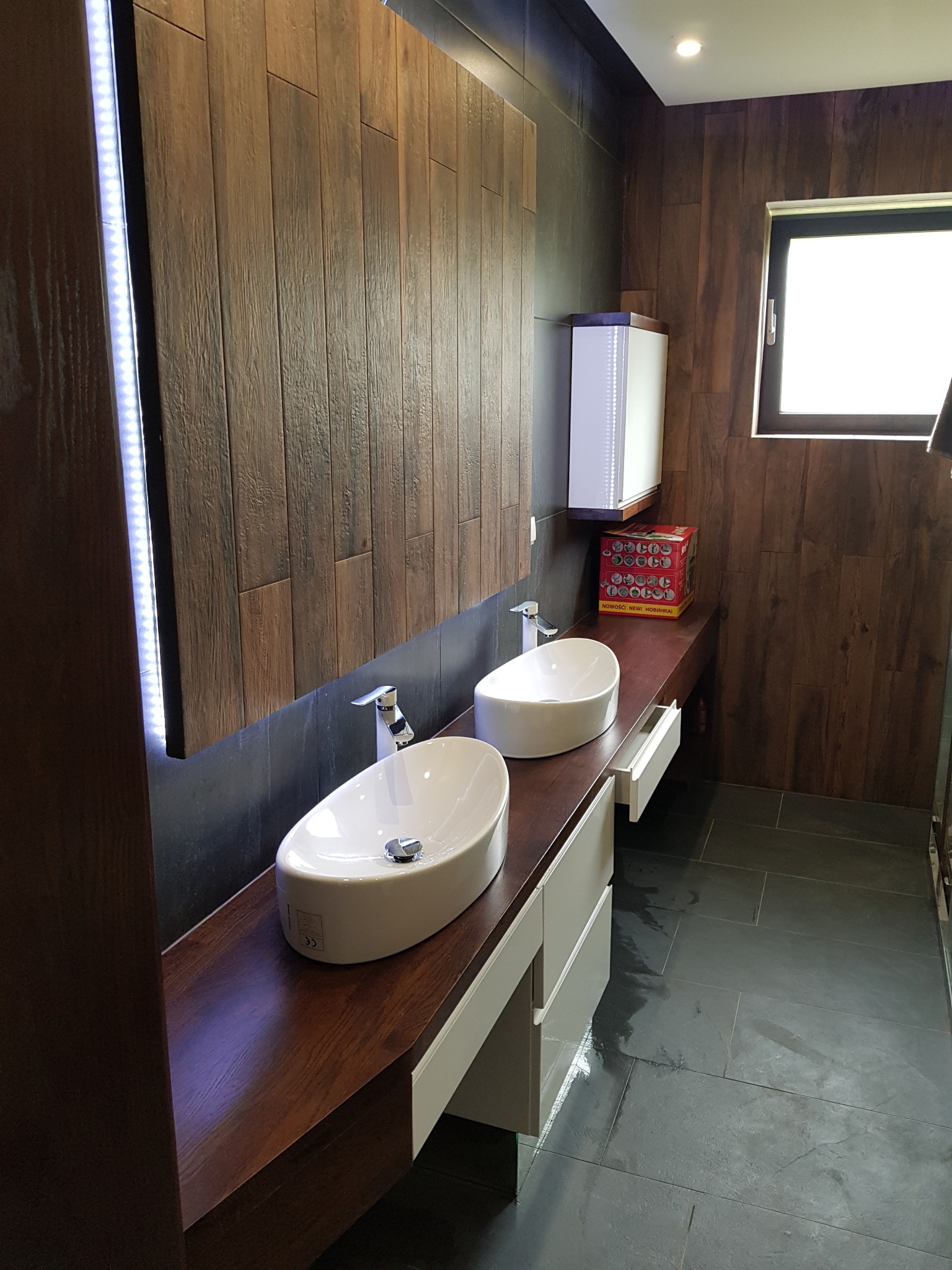 Nowoczesna łazienka z dwoma owalnymi umywalkami nablatowymi na blacie z ciemnego drewna, podświetlane lustro LED, ściany wyłożone płytkami imitującymi drewno, szare płytki podłogowe.