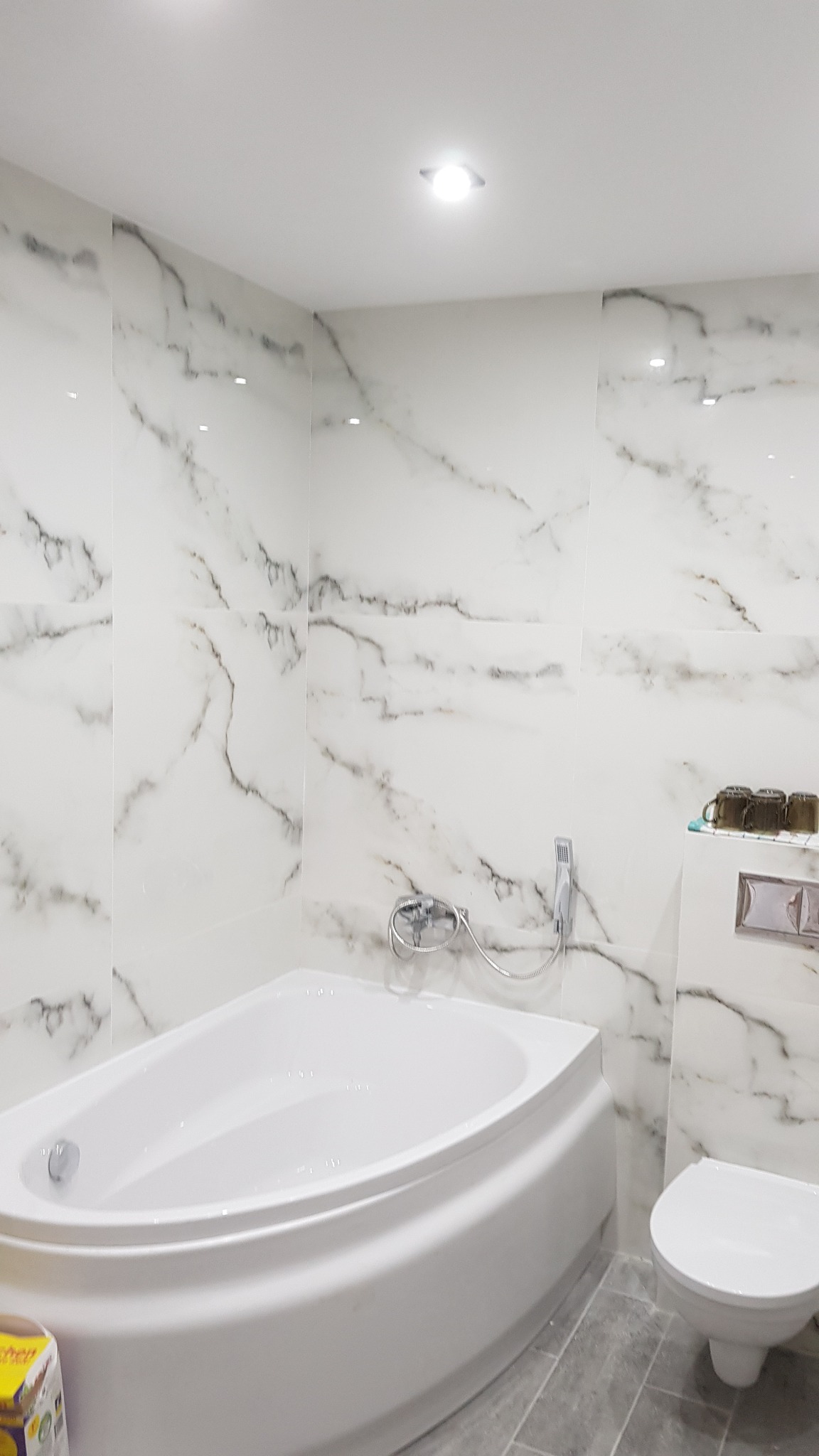 Nowoczesna łazienka z białą wanną narożną, toaletą i marmurowymi płytkami ściennymi z szarymi żyłami, oświetlona punktowo.