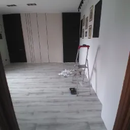 Pokój po remoncie z jasnymi panelami podłogowymi, białe ściany z czarnymi obrazami i listwą oświetleniową, aluminiowa drabina, narzędzia malarskie i białe rozety sufitowe.