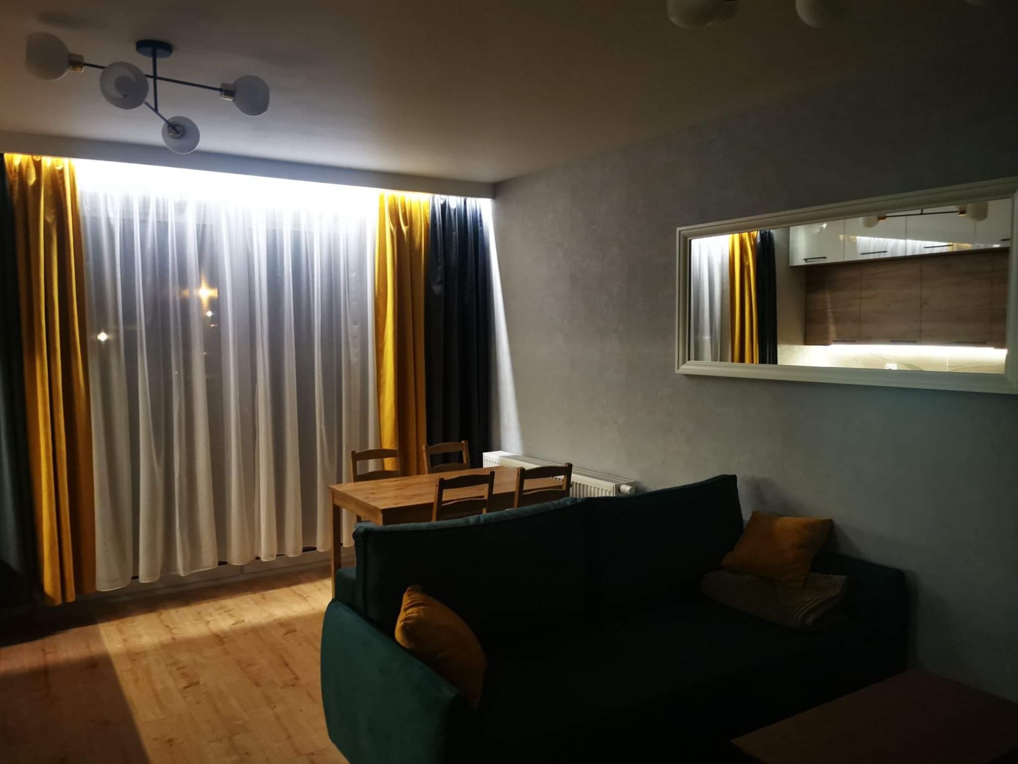 Pokój dzienny z drewnianą podłogą, zieloną kanapą z pomarańczowymi poduszkami, drewnianym stołem z krzesłami i oknem z białymi, żółtymi i czarnymi zasłonami. W lustrze odbija się część kuchni...