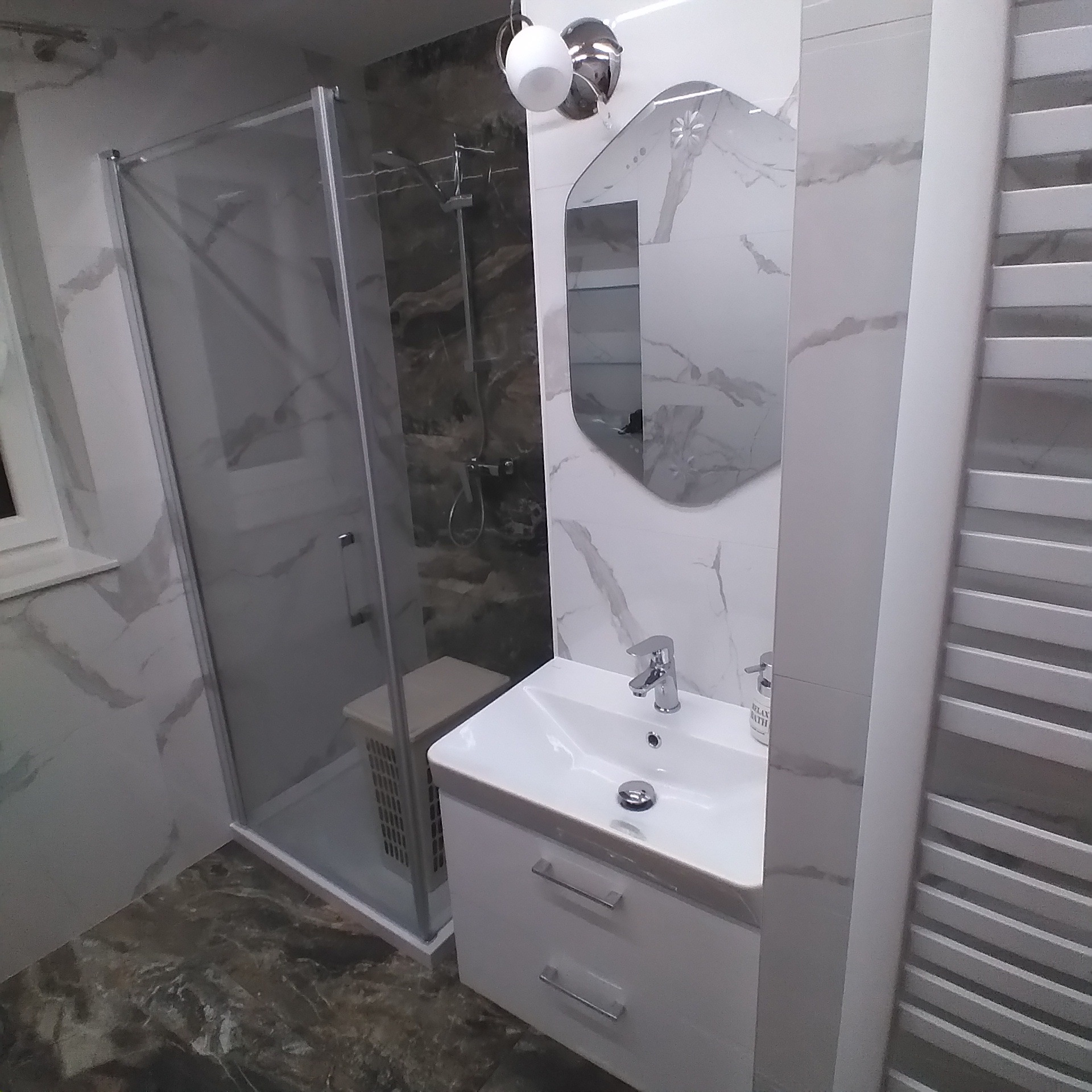 Nowoczesna łazienka z kabiną prysznicową, umywalką z szafką i lustrem o nieregularnym kształcie, wykończona płytkami imitującymi marmur.