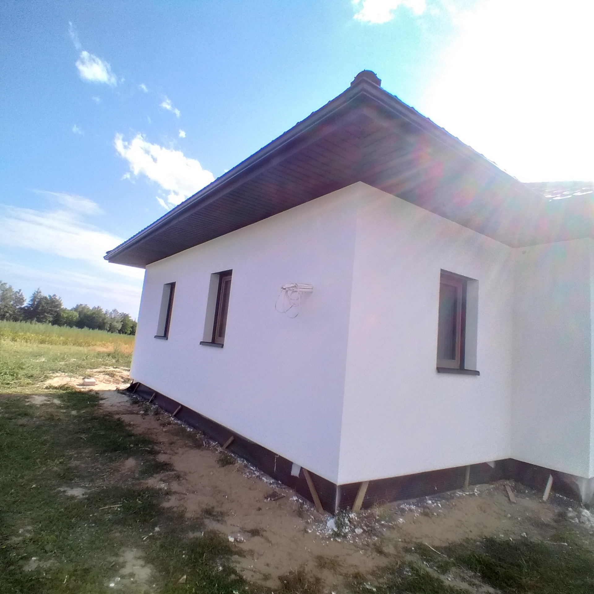 Boczny widok na otynkowany dom jednorodzinny z brązowymi oknami, widoczne elementy instalacji na ścianie, częściowo zagospodarowany teren wokół budynku.