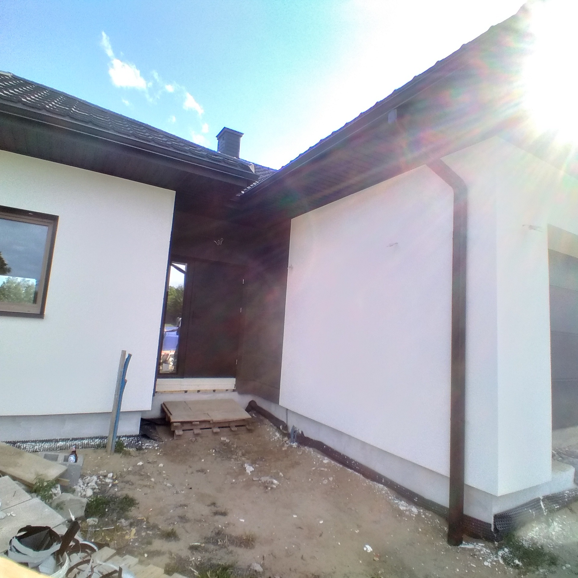 Nowo otynkowana, biała elewacja domu jednorodzinnego z ciemnymi rynnami i podbitką, widok na narożnik budynku w słoneczny dzień.