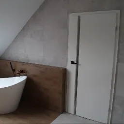 Nowoczesna łazienka na poddaszu z wolnostojącą wanną, baterią w kolorze miedzi i drewnianą zabudową przy wannie oraz białą stolarką drzwiową i szarą płytką.