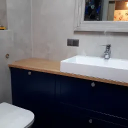 Nowoczesna łazienka z umywalką nablatową na szafce w odcieniach granatu, drewniany blat, szara ściana z widocznym włącznikiem światła i lustro z odbiciem obrazka z odciskami dłoni.