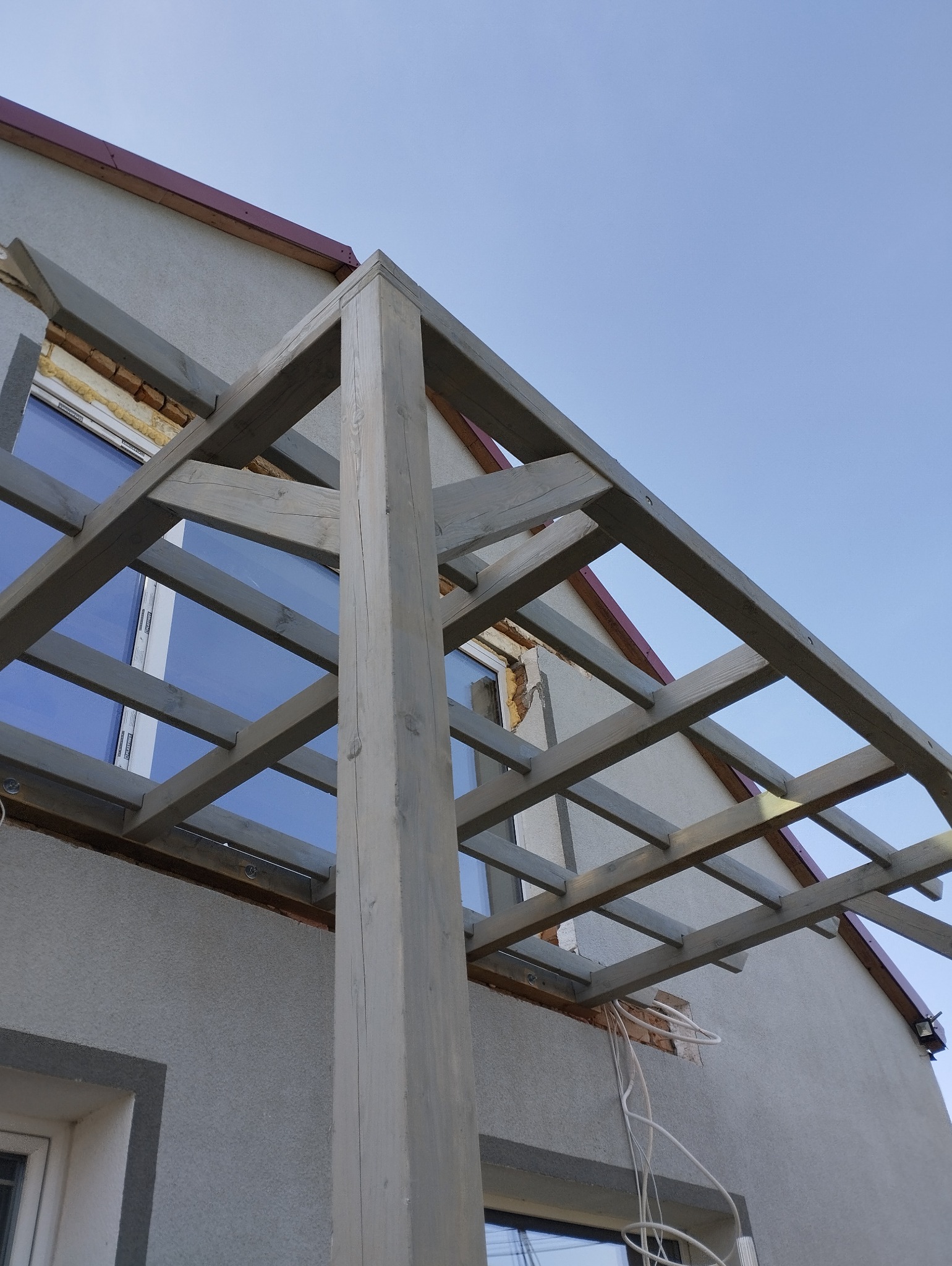 Drewniana konstrukcja z belek i słupów, prawdopodobnie zadaszenie tarasu, w trakcie budowy przy szarej elewacji budynku z widocznymi przewodami elektrycznymi.
