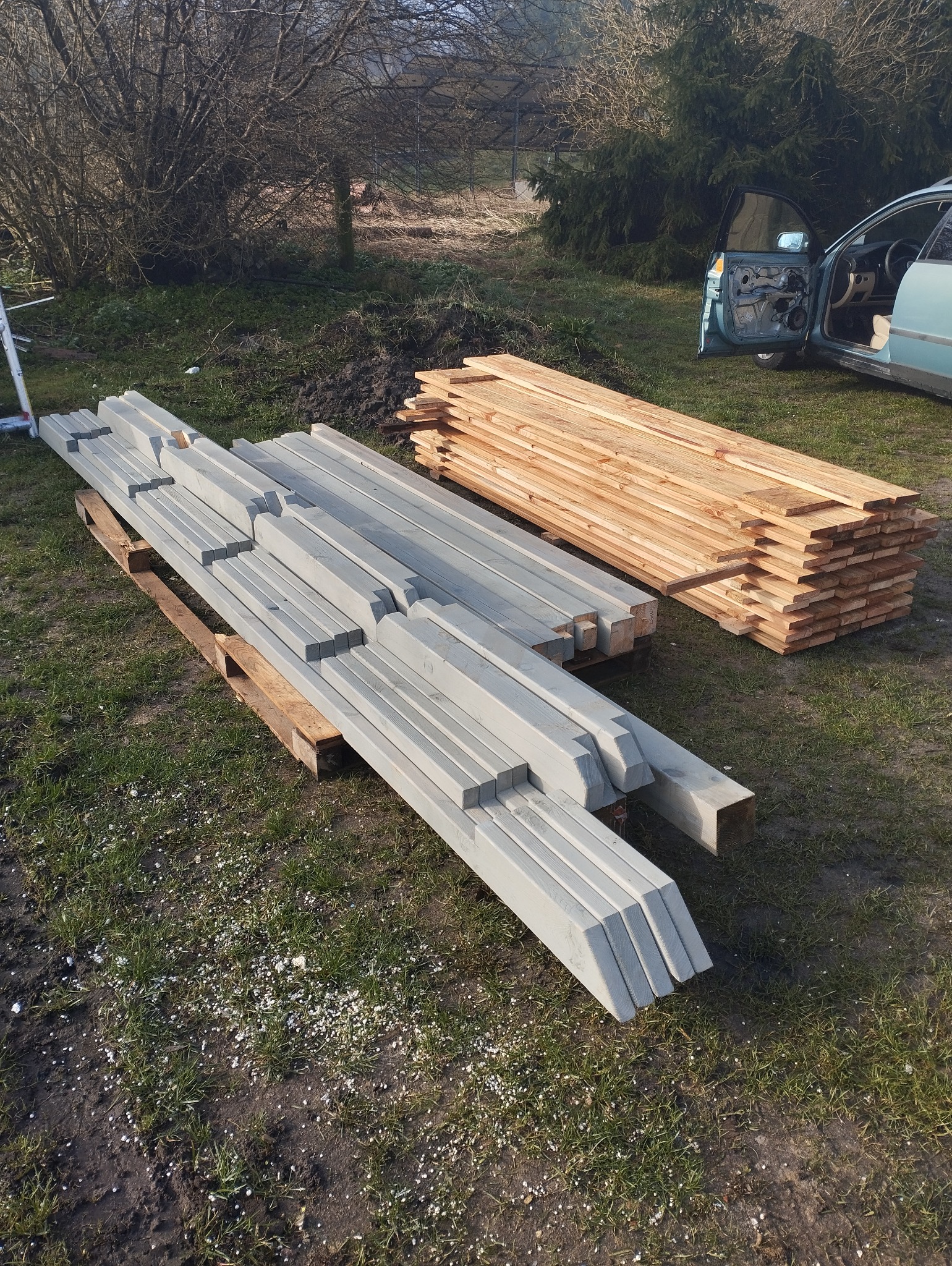 Przygotowane elementy konstrukcyjne z drewna: szare kantówki na palecie i deski, na zewnątrz, w tle samochód z otwartymi drzwiami.