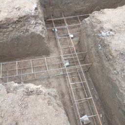 Mk bud - Zbrojenie fundamentów w wykopie, widok z góry na stalową siatkę zbrojeniową ułożoną w przygotowanych rowach w ziemi.