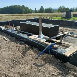 Przygotowanie do wylewki fundamentów: widoczne zbrojenie, izolacja z folii kubełkowej i szalunki na gruncie z butelką wody.