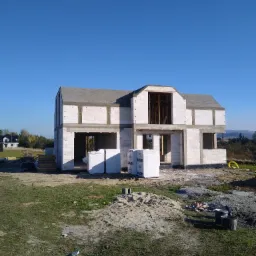 Dwukondygnacyjny dom murowany w stanie surowym otwartym, z widocznymi paletami bloczków SOLBET, na tle błękitnego nieba i odległego krajobrazu.