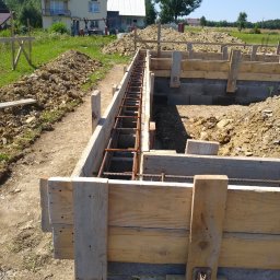 Mk bud - Przygotowanie szalunku i zbrojenia pod fundament na tle domu jednorodzinnego w słoneczny dzień.