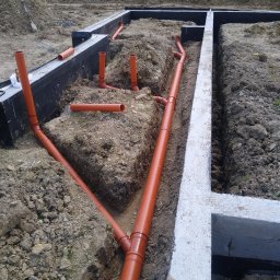 Mk bud - Wykop pod fundamenty z ułożonymi rurami kanalizacyjnymi w ziemi, zabezpieczone czarną folią, widok z góry.