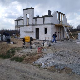 Mk bud - Wznoszenie dwukondygnacyjnego budynku z białych bloczków, widoczna żółta betoniarka i pracownik w niebieskim kombinezonie.