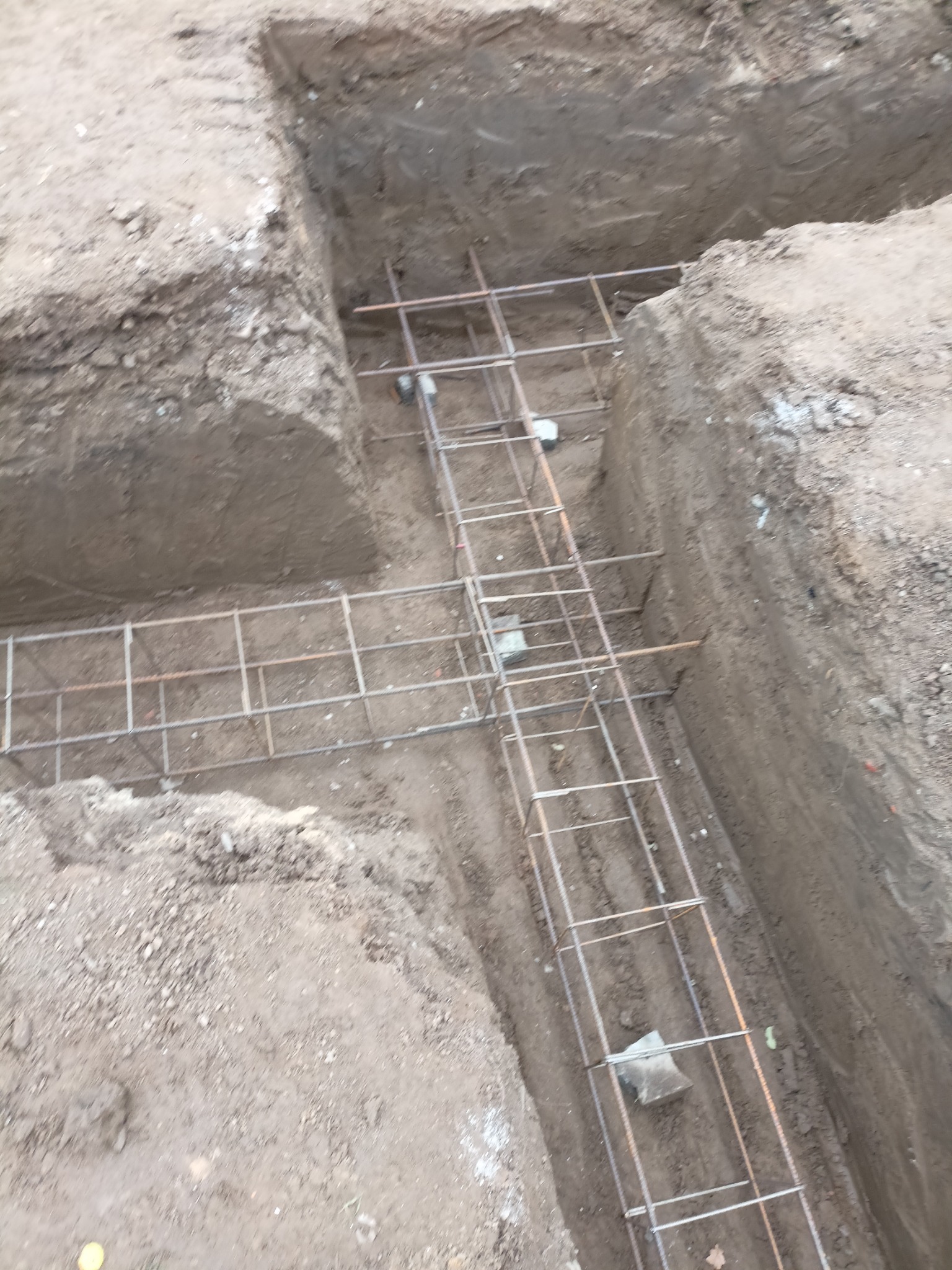 Zbrojenie fundamentów w wykopie, widok z góry na stalową siatkę zbrojeniową ułożoną w przygotowanych rowach w ziemi.