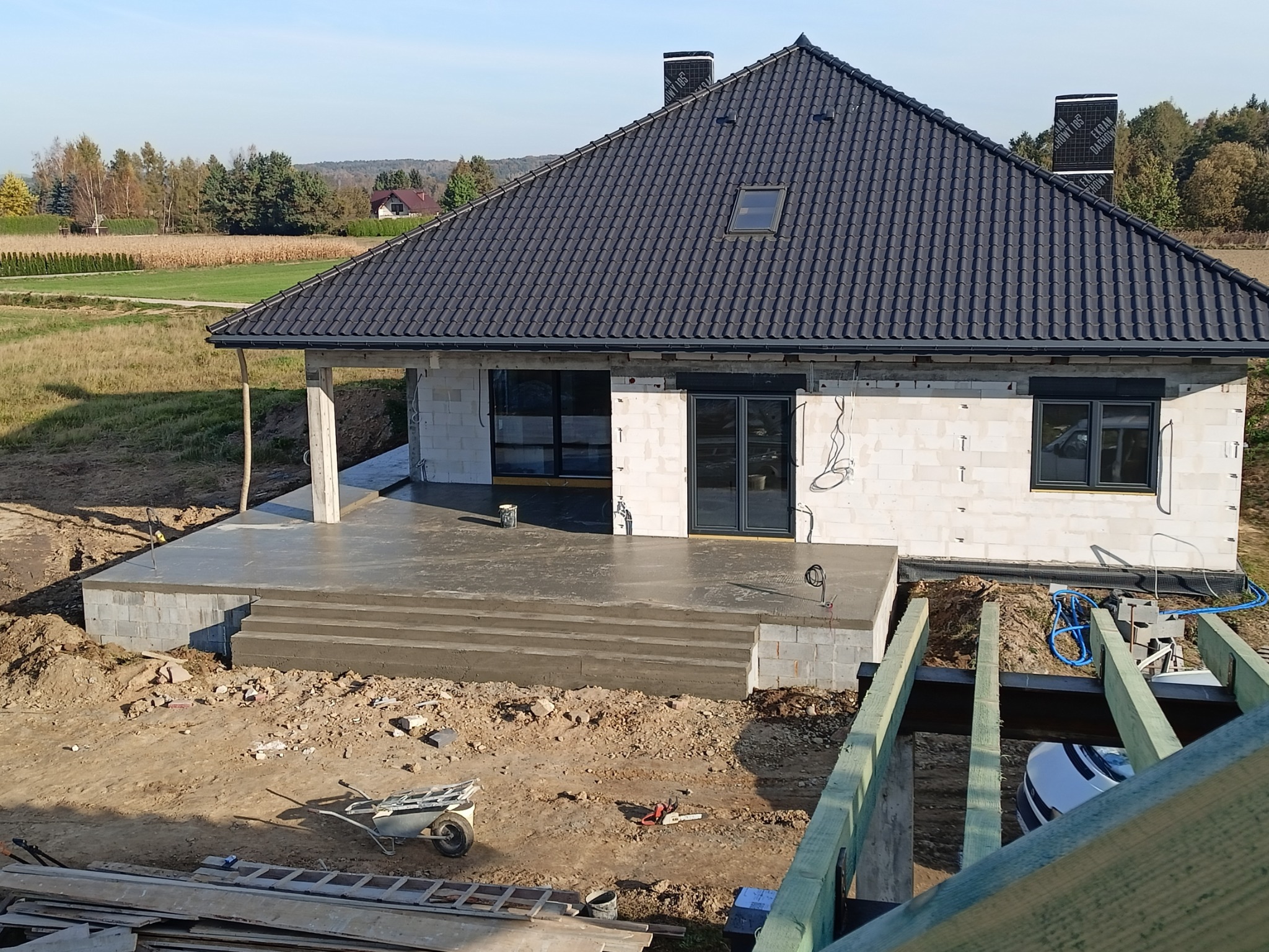 Surowy stan budowy domu jednorodzinnego z czarną dachówką, widoczne fundamenty tarasu z betonowymi schodami, ściany z bloczków, częściowo zamontowane okna i drzwi, oraz widok na okoliczne pola...