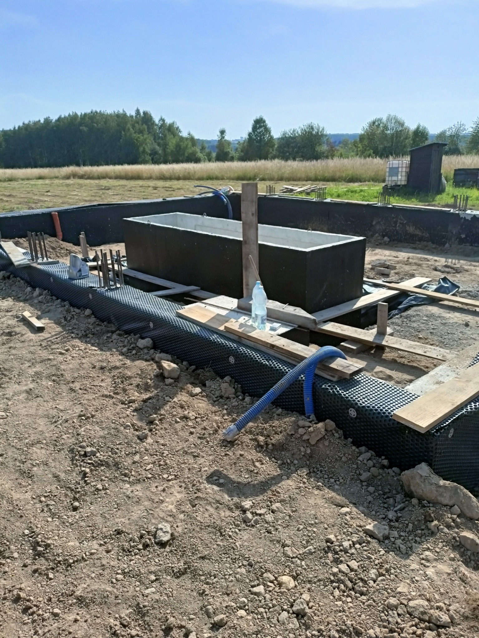 Przygotowanie do wylewki fundamentów: widoczne zbrojenie, izolacja z folii kubełkowej i szalunki na gruncie z butelką wody.