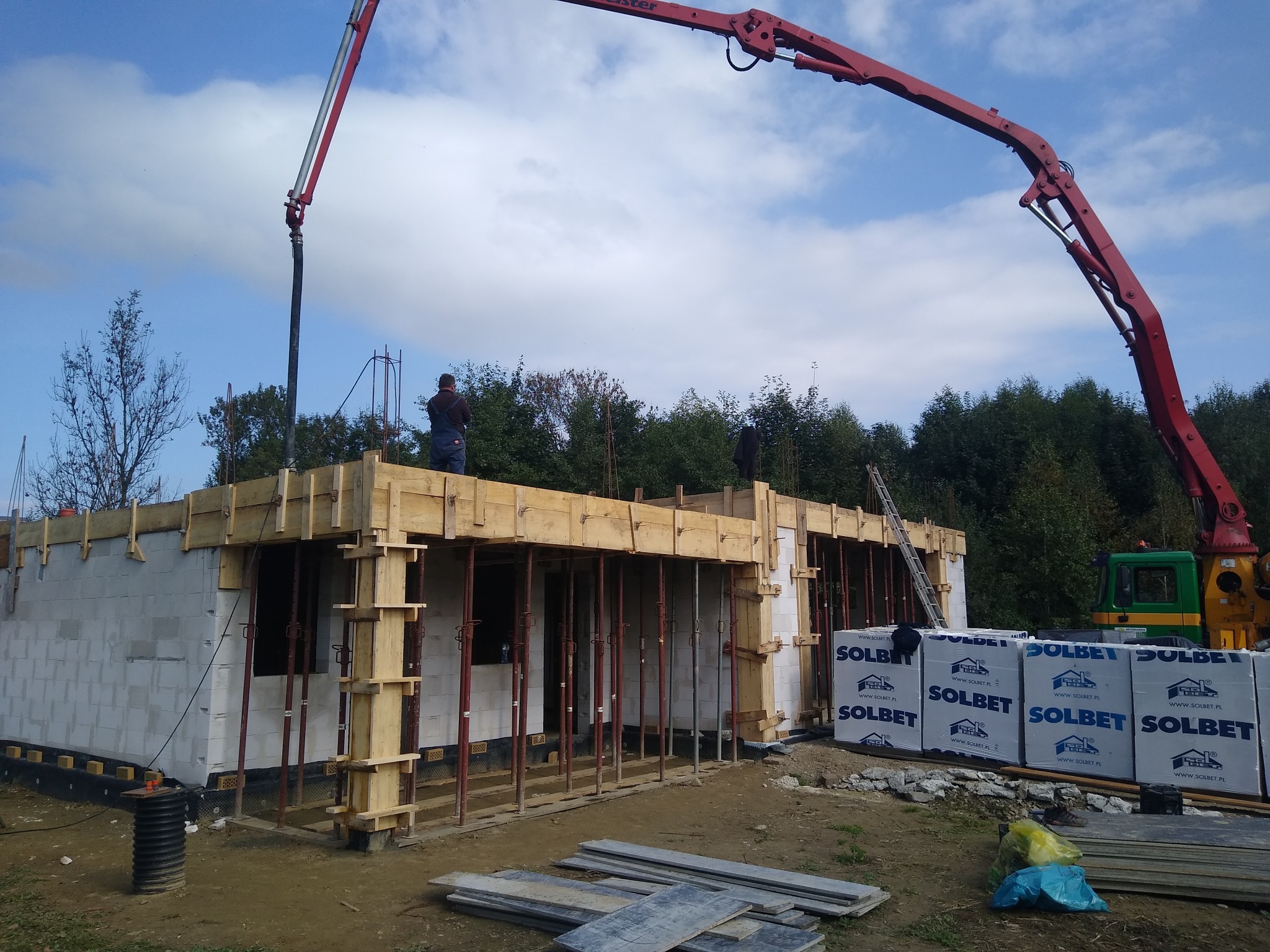 Wylewanie betonu na strop domu w trakcie budowy z użyciem pompy do betonu, widoczne szalunki i pracownicy na budowie, palety z bloczkami Solbet