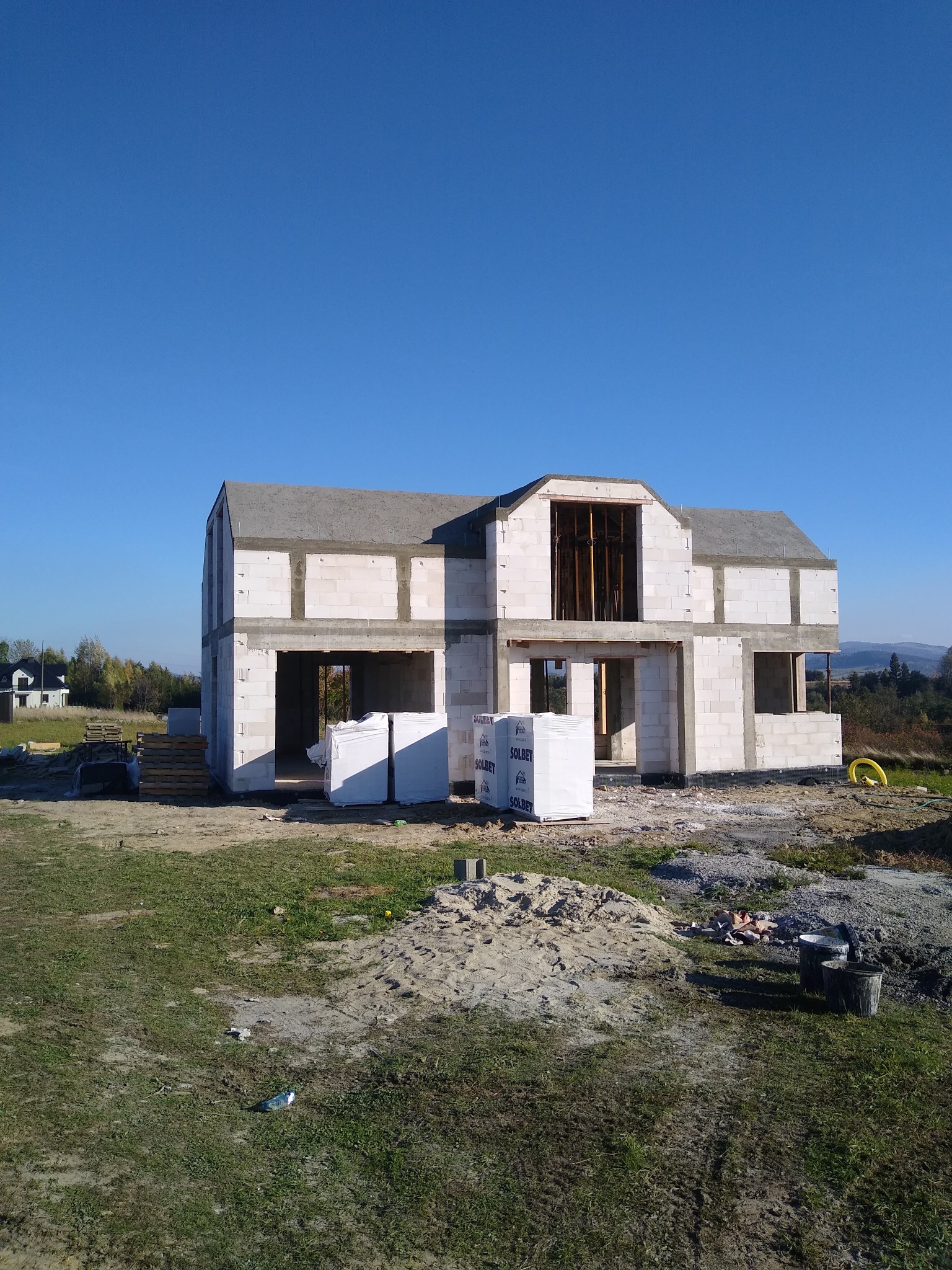 Dwukondygnacyjny dom murowany w stanie surowym otwartym, z widocznymi paletami bloczków SOLBET, na tle błękitnego nieba i odległego krajobrazu.