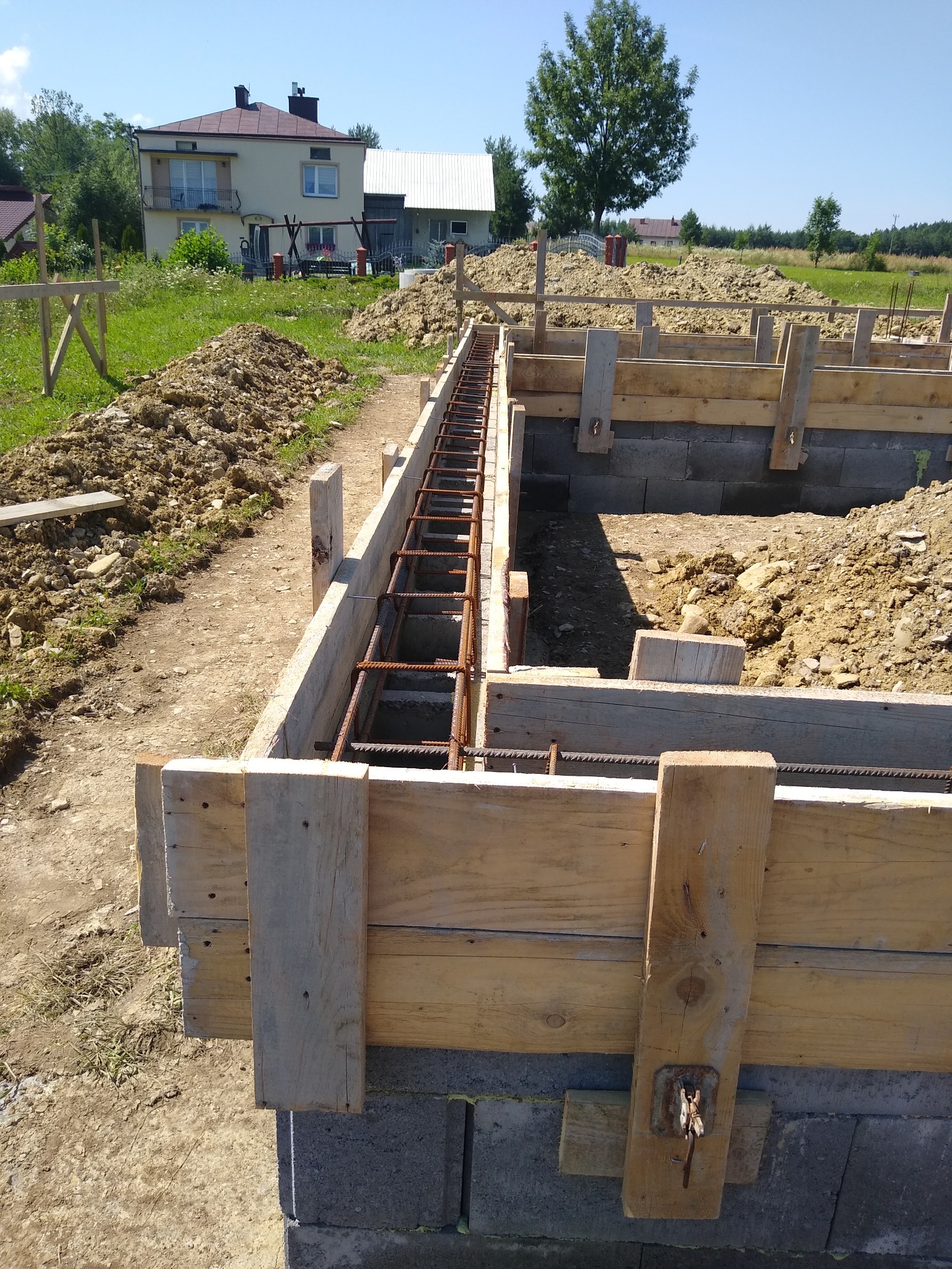 Przygotowanie szalunku i zbrojenia pod fundament na tle domu jednorodzinnego w słoneczny dzień.