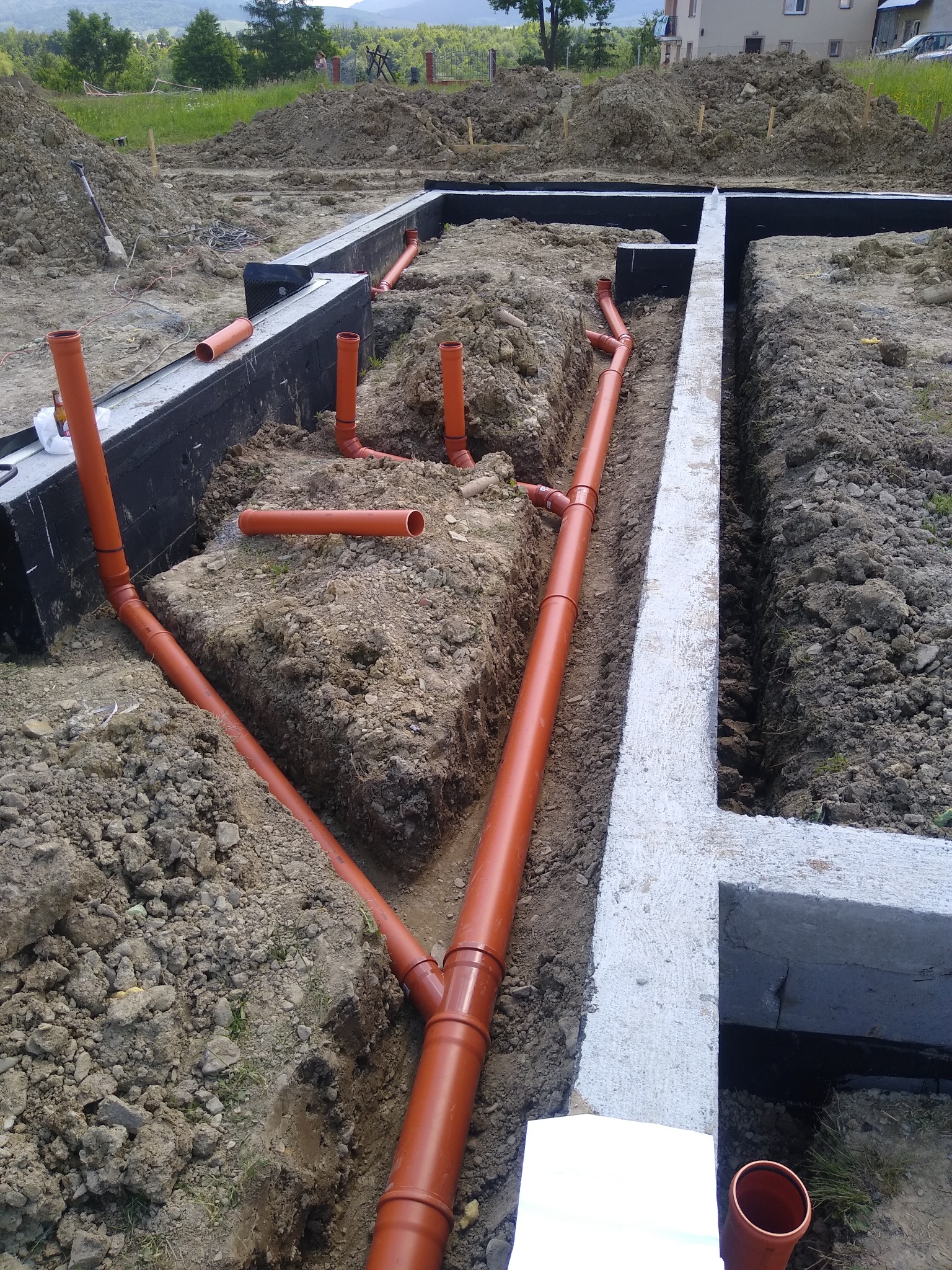 Wykop pod fundamenty z ułożonymi rurami kanalizacyjnymi w ziemi, zabezpieczone czarną folią, widok z góry.