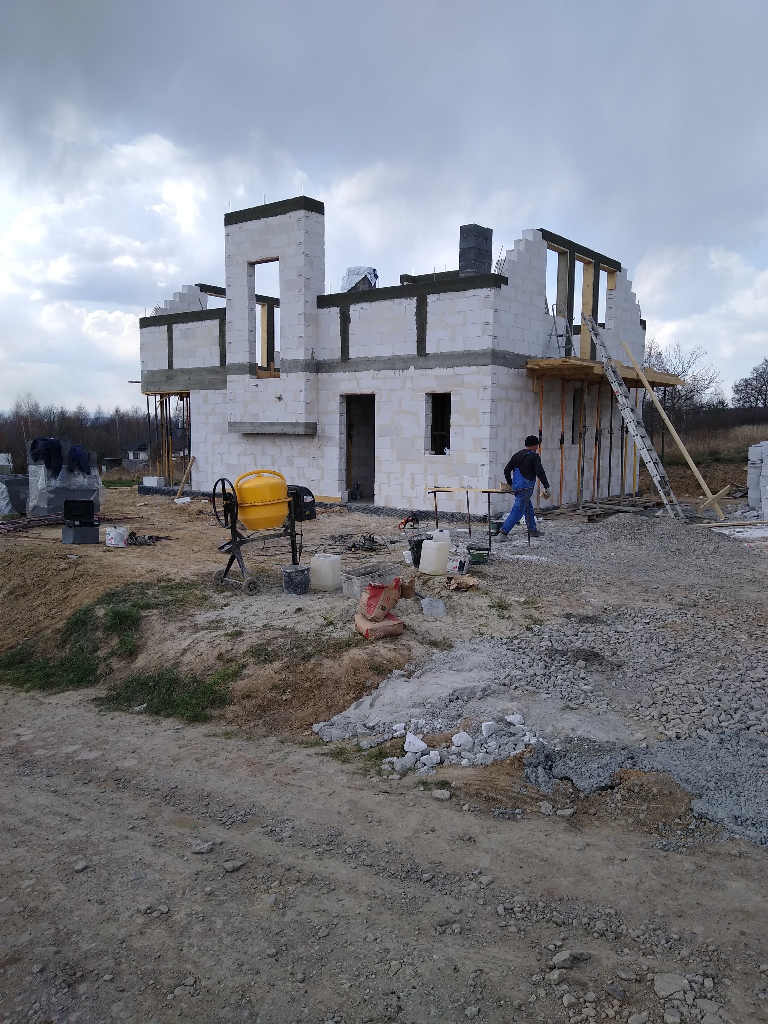 Wznoszenie dwukondygnacyjnego budynku z białych bloczków, widoczna żółta betoniarka i pracownik w niebieskim kombinezonie.