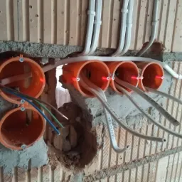 Montaż przewodów elektrycznych w pomarańczowych puszkach instalacyjnych w surowej ścianie z cegły, widoczna wiązka przewodów z laserowym poziomowaniem.