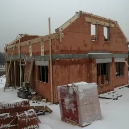 Surowy stan domu z czerwonej cegły ceramicznej Porotherm w zimowej scenerii, widoczne drewniane elementy konstrukcyjne i zapas materiałów budowlanych na śniegu, szary dzień.