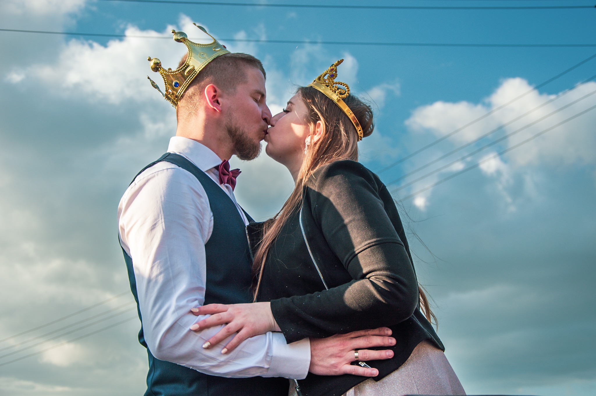 Para w koronach, on w kamizelce i muszce, ona w czarnej kurtce, całująca się na tle pochmurnego nieba z liniami energetycznymi.