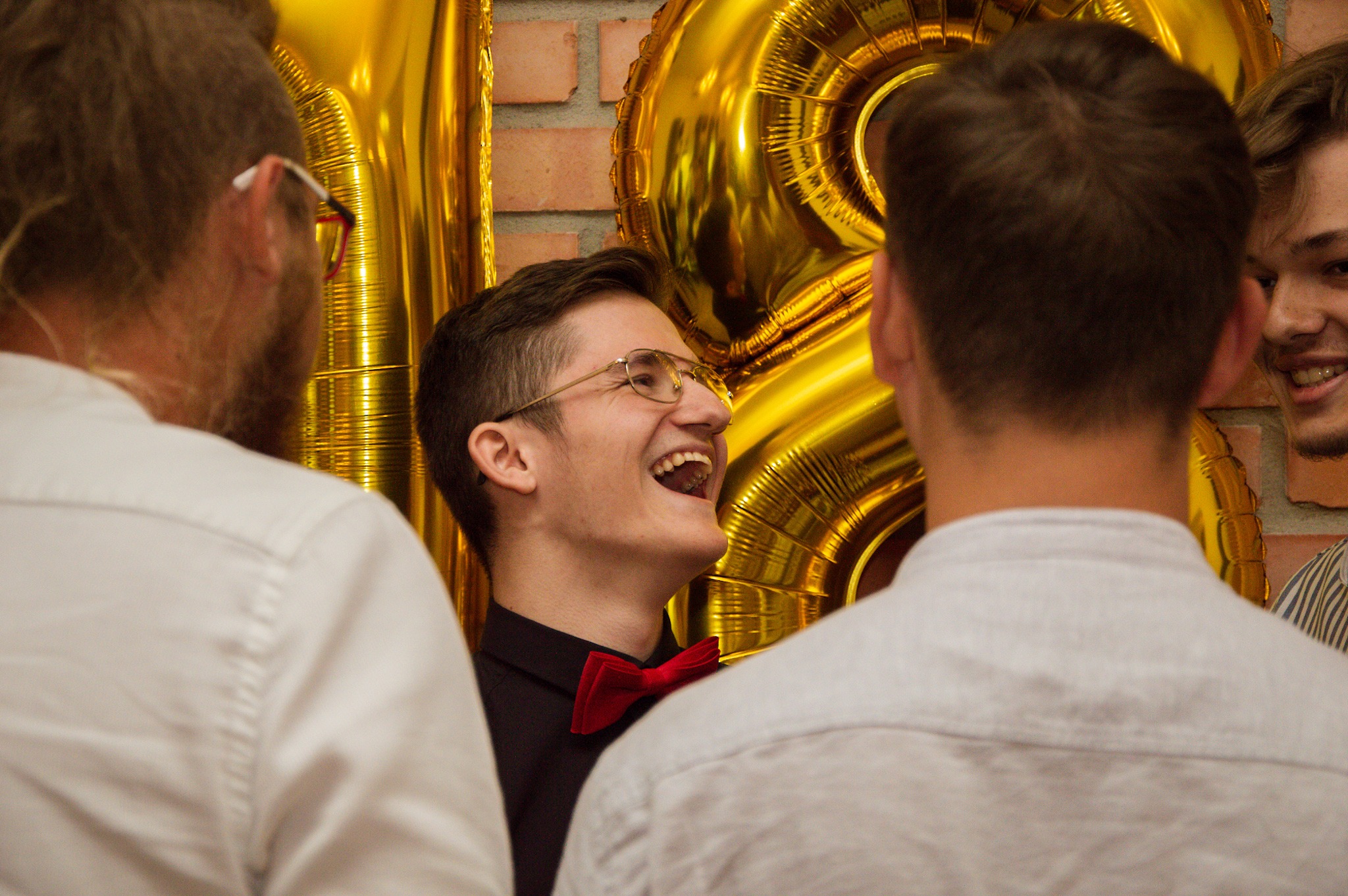 Rozesmiany mężczyzna w okularach i czerwonej muszce, otoczony przez inne osoby, na tle złotych balonów cyfr podczas wydarzenia.